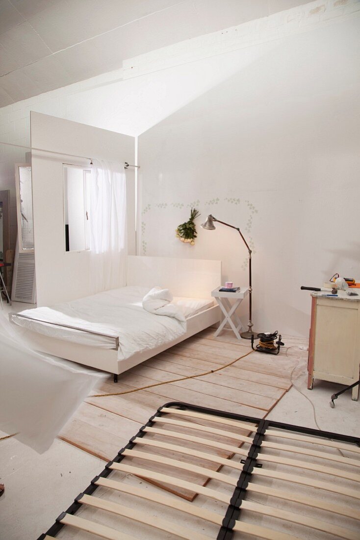 Slated bed frame on floor; white double bed and standard lamp in background