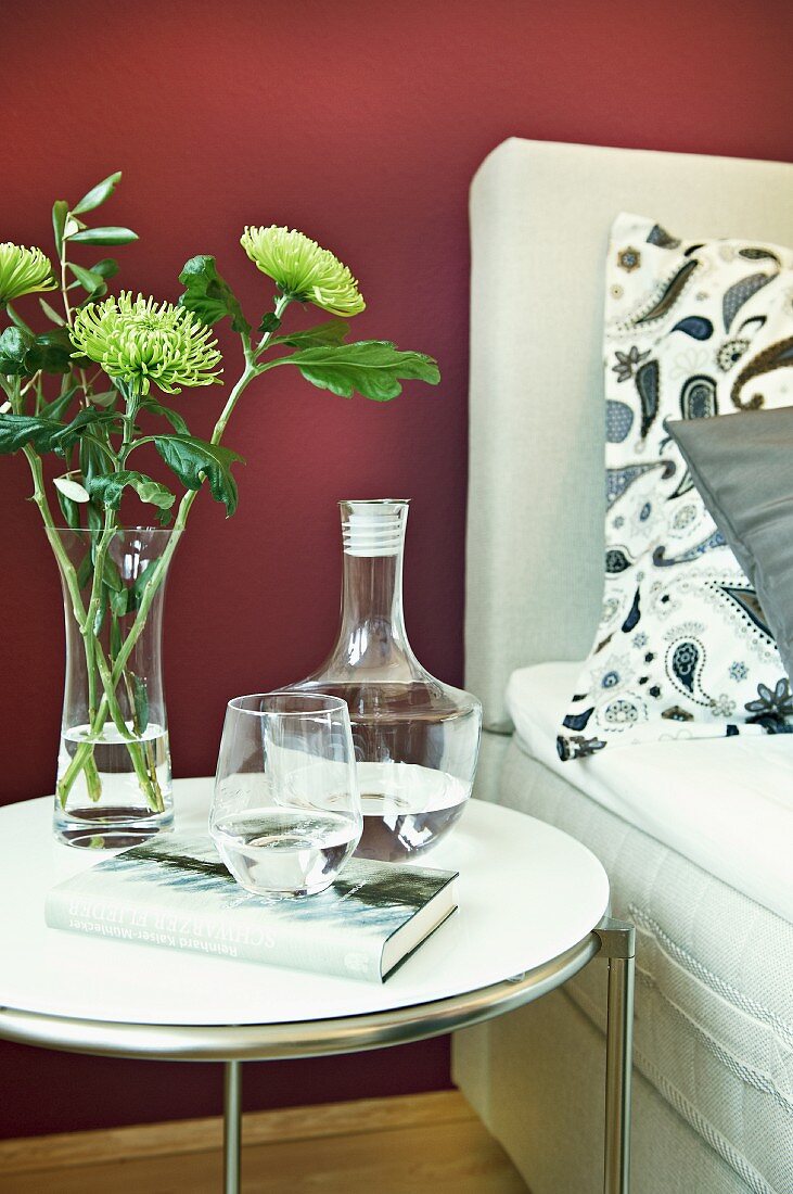A carafe of water with a drinking glass and a bunch of flowers on a bedside table against a purple wall