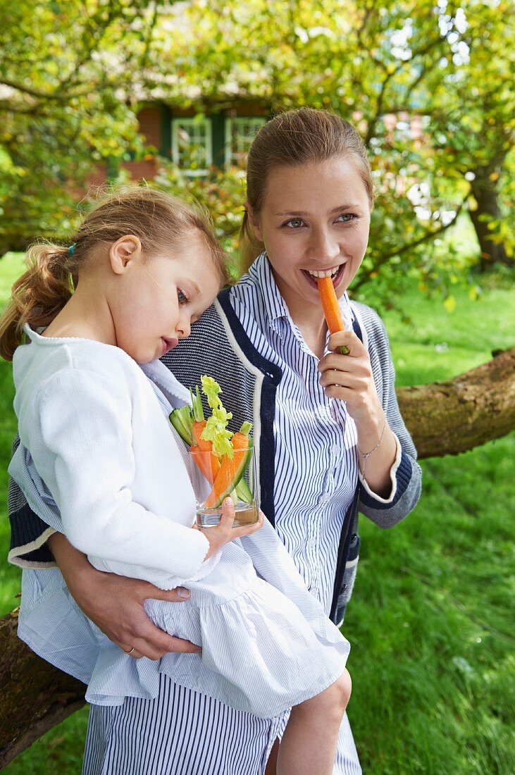 Frau mit kleinem Mädchen isst Gemüsesticks im Garten