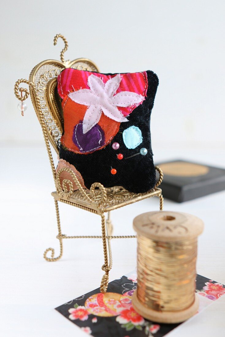 Hand-sewn, velvet pincushion with floral motif on miniature chair next to reel of gold thread