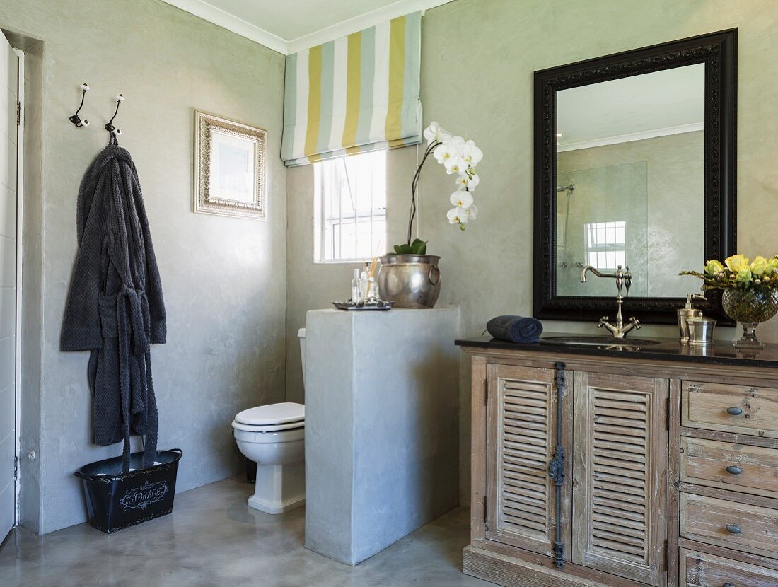 Toilet screened by half-height wall next to vintage, mango-wood washstand below black-framed mirror