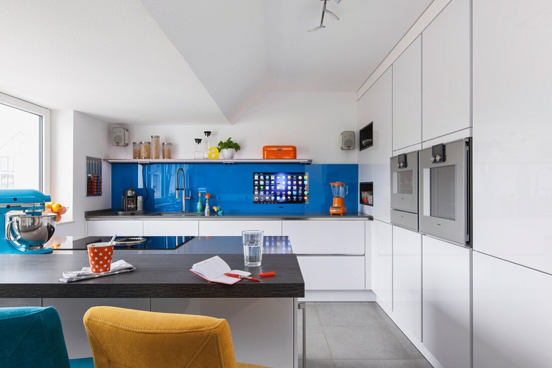 A breakfast bar in a white designer kitchen with a blue glass panel on the wall with a built-in touch screen