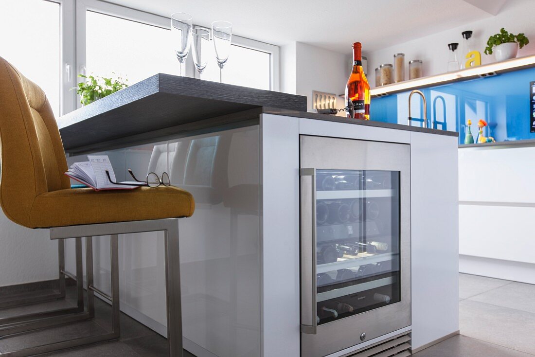 A kitchen island with an integrated wine fridge and a yellow upholstered stool in a designer kitchen