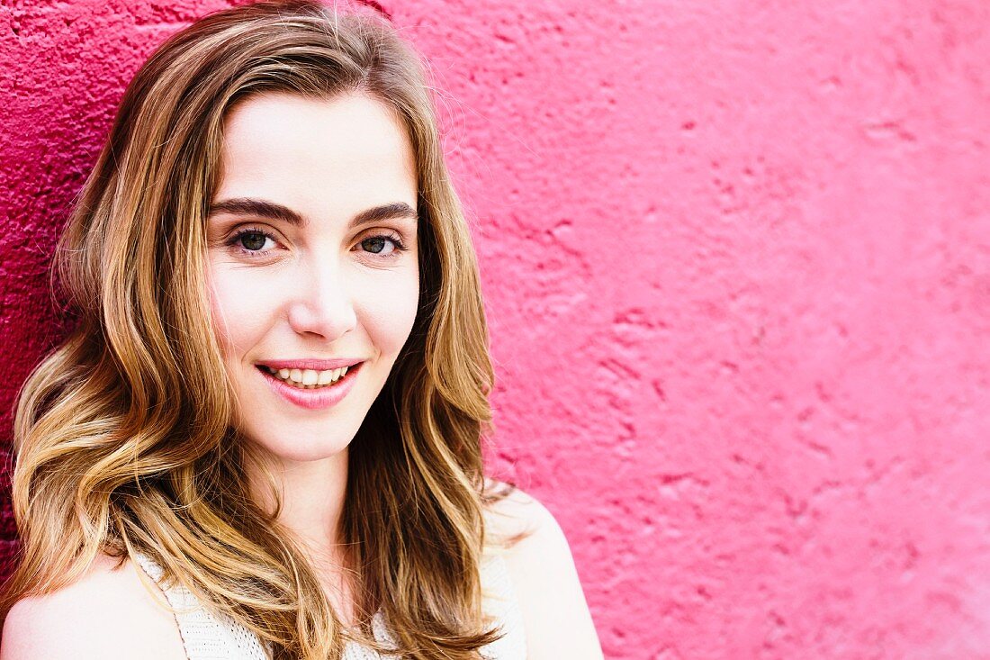 A portrait of a dark blonde woman against a pink wall