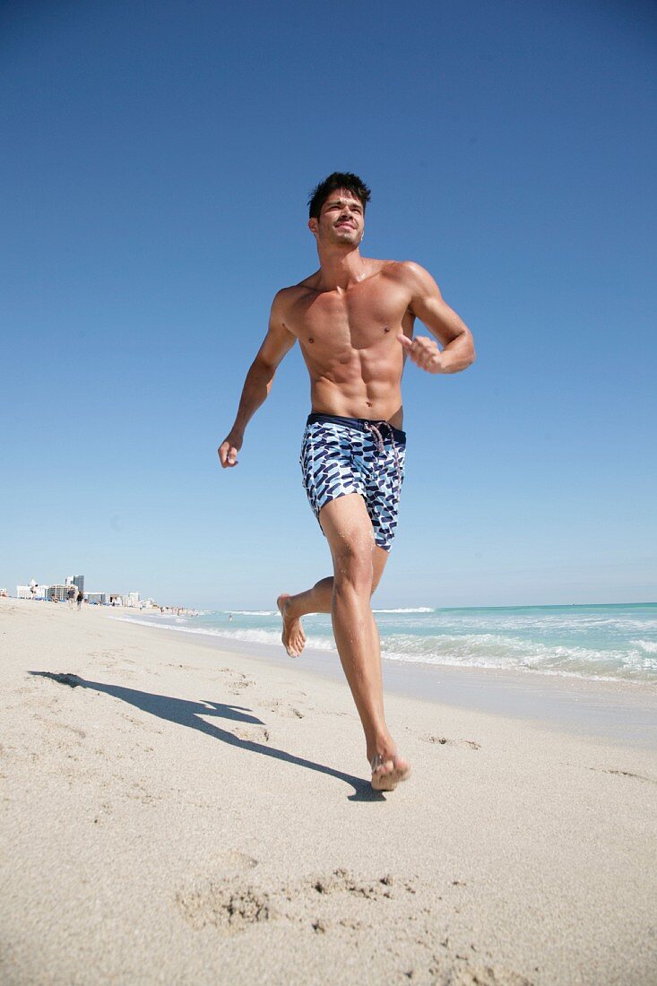 A young, topless man wearing shorts jogging along a beach