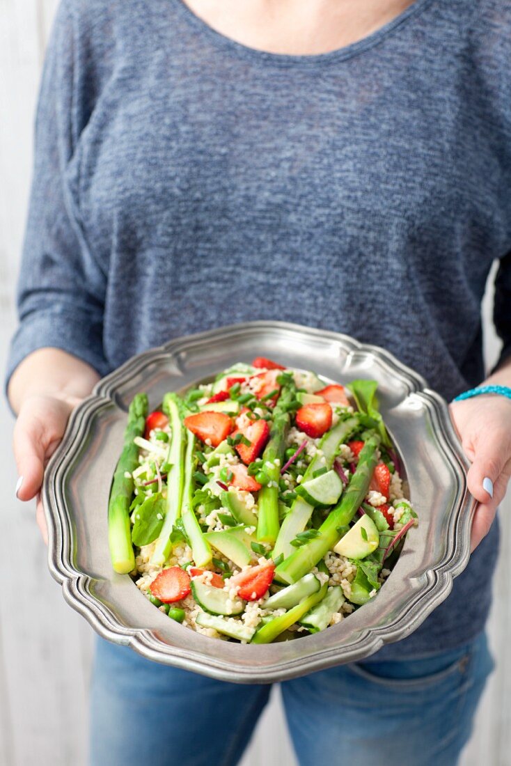 A woman serving bulgur and asparagus salad with strawberries, peas and young beetroot leaves