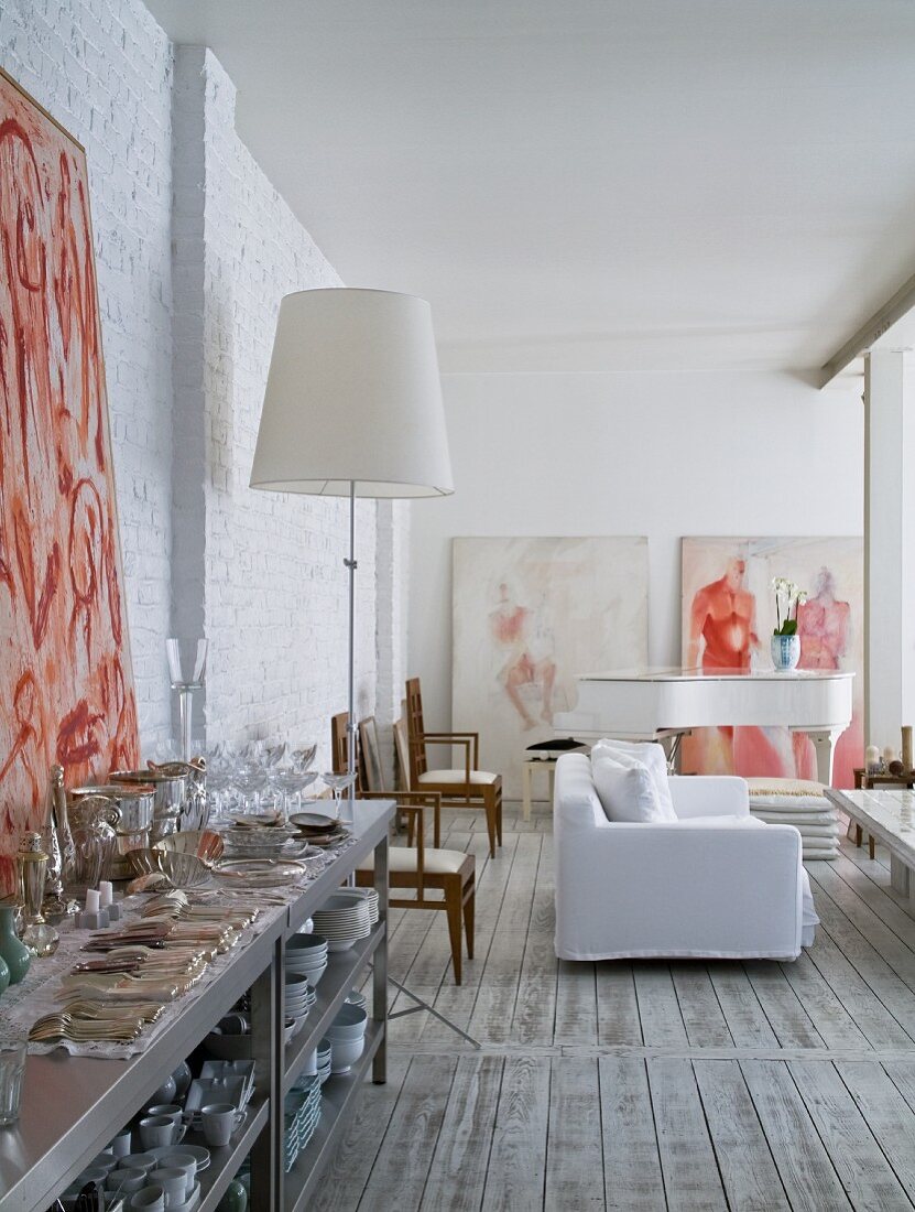 Cutlery and crockery on stainless steel shelving and white loose-covered sofa and grand piano in background of open-plan interior with whitewashed brick walls