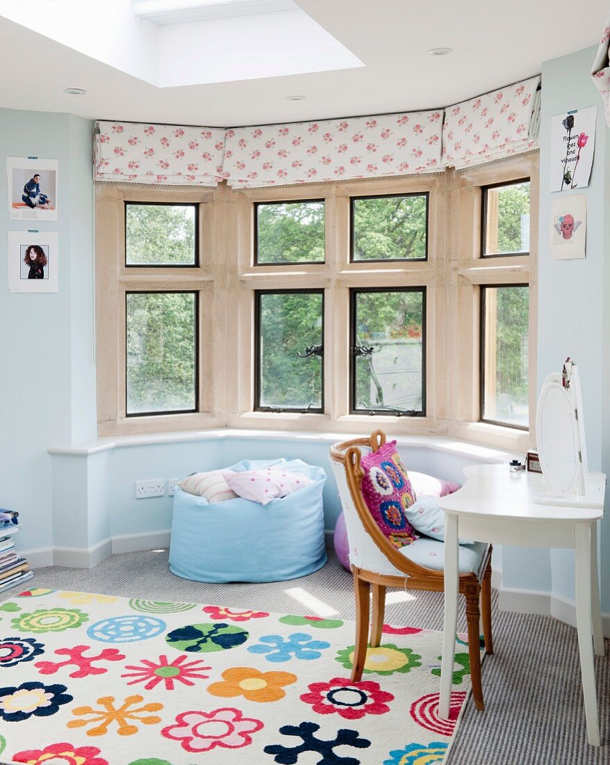 Brightly patterned rug in front of comfortable beanbag in window bay and antique chair at white table to one side in teenager's bedroom