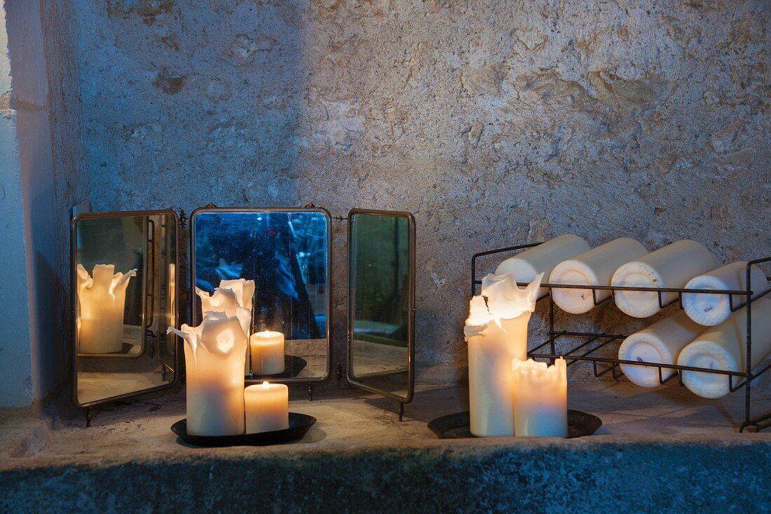 Brennende Stumpenkerzen und kleiner dreiteiliger Spiegel neben Metallgestell mit Kerzen auf Steinablage