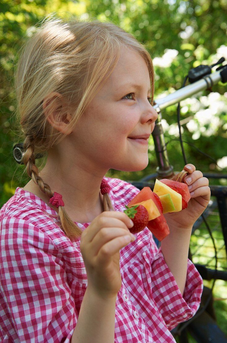 A little girl in a garden holding a fruit skewer