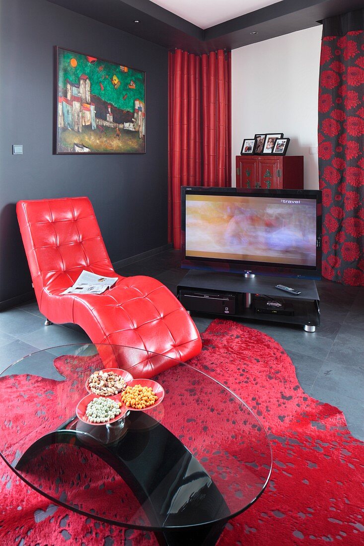 Glass coffee table in front of easy chair with red leather cover; TV on floor in background
