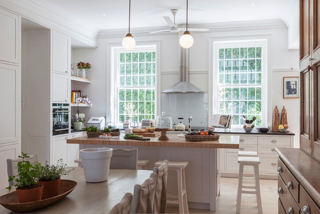 Blick über Essplatz auf freistehende Kücheninsel in Landhausküche mit weissen Schrankfronten, im Hintergrund Sprossenfenster