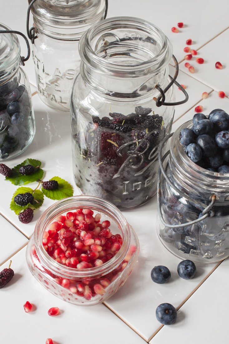 Fresh blueberries, mulberries and pomegranate seeds in preserving jars on white tiles