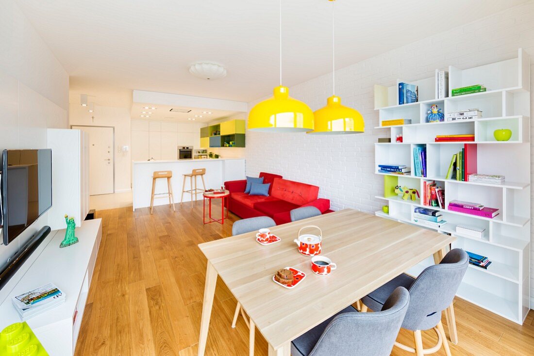 Dining area with yellow pendant lamps in front of bookcase, media centre, red sofa and open-plan kitchen with breakfast bar