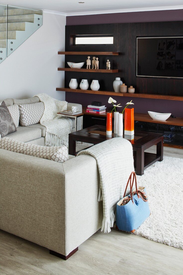 Elegant lounge area with pale sofa set and collection of vases on coffee table opposite wall painted dark purple with wooden shelves