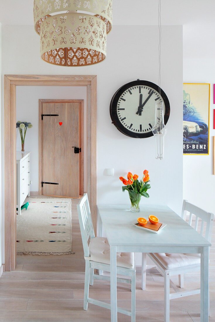 Station clock above small table with two chairs; section of pendant lamp in foreground and doorway leading to hall in background