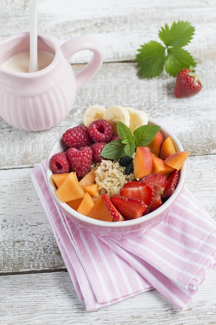 A bowl of quinoa, fresh fruit and mint leaves, a jug of milk and a strawberry with a leaf
