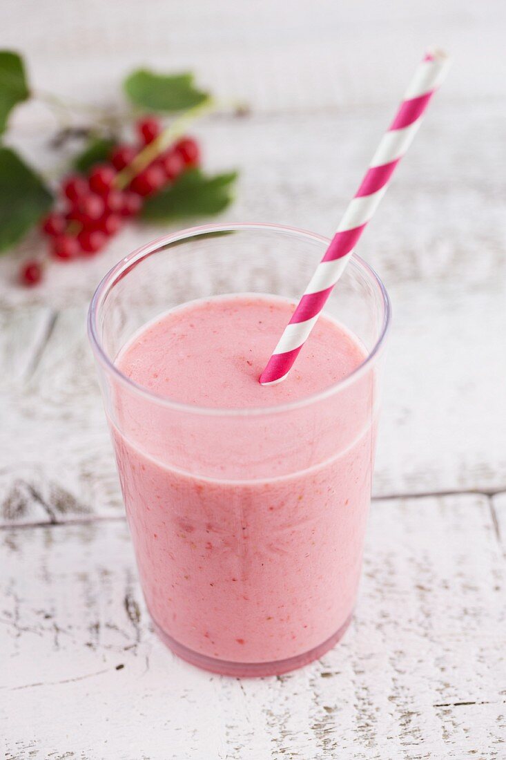 A redcurrant smoothies in a glass with a paper straw and a sprig on redcurrants in the background