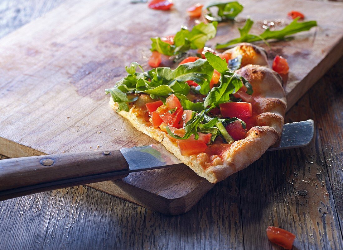 A slice of pizza bread with tomatoes and rocket on a wooden board