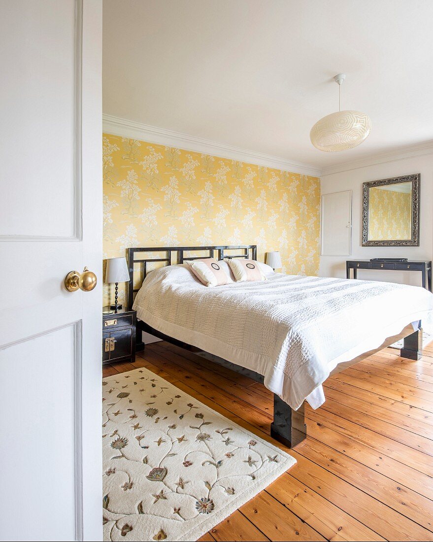 Double bed against accent wall with yellow wallpaper in simple master bedroom with rustic wooden floor seen through open door