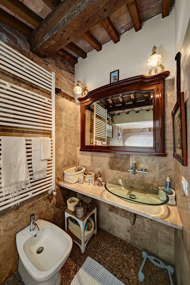 Rustic bathroom with wood-beamed ceiling, washstand with modern glass basin and white, heated towel rail above bidet to one side