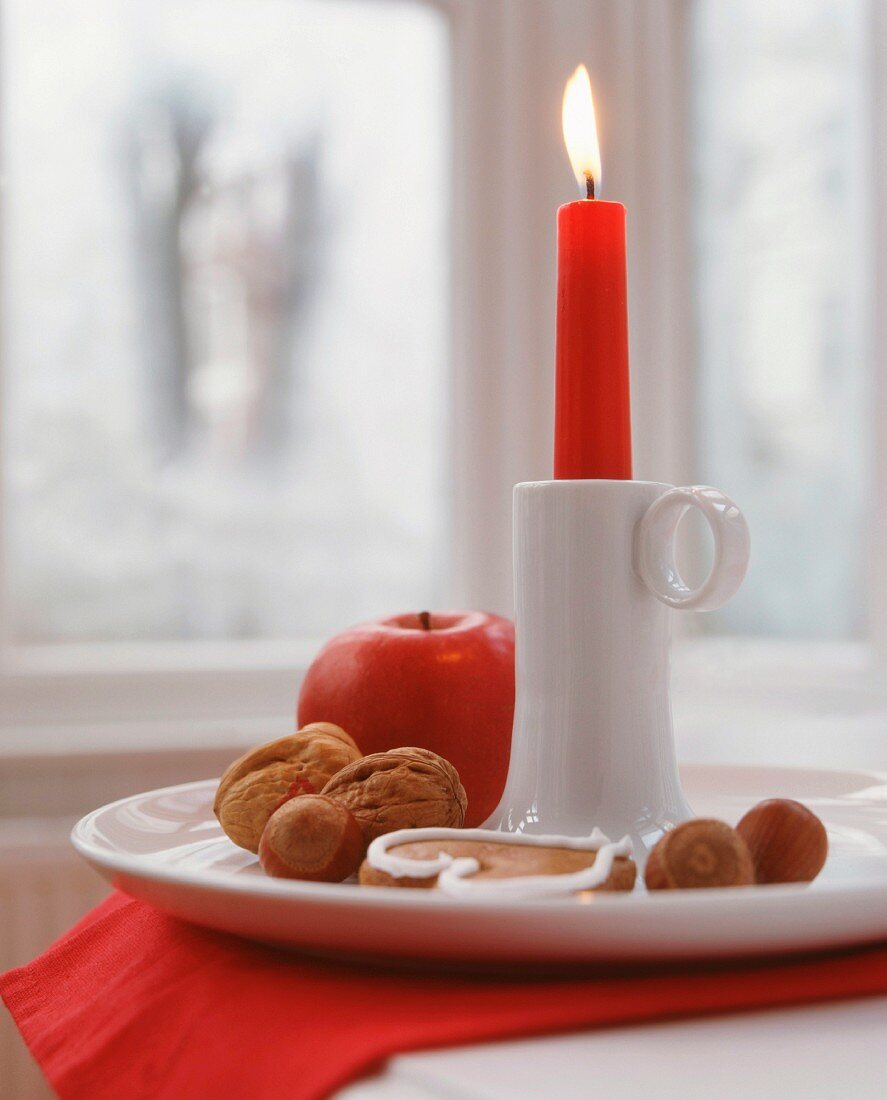 Brennende rote Kerze in weißer Kerzenständer aus Porzellan mit Nüssen, Apfel und Lebkuchen