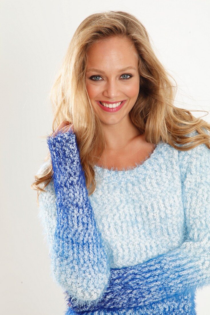 A blonde woman wearing a blue-toned, coarse-knit jumper with her hand on her hair