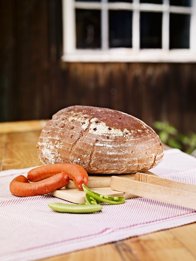 A whole loaf of wholemeal rye bread, two sausages and pea pods on a table