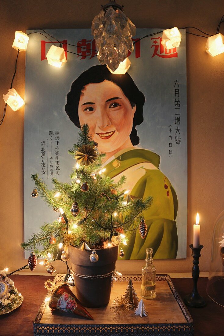 Small, decorated Christmas tree with fairy lights in front of illuminated poster of Japanese woman