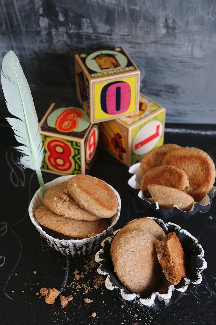 Kekse aus Mandelmehl und Honig in handgemachten Tonschälchen, Spielzeugwürfel und Feder, auf schwarzem Untergrund