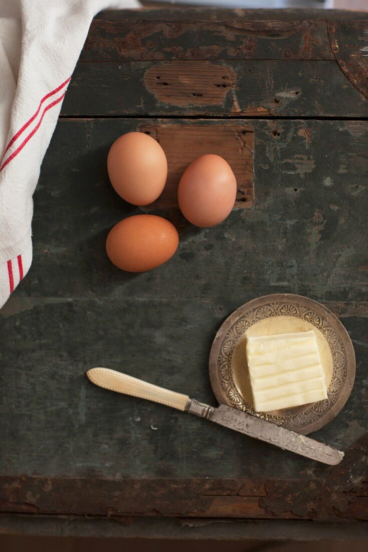 Rustikales Stillleben mit Eiern und Butter mit Messer auf Holztisch (Aufsicht)