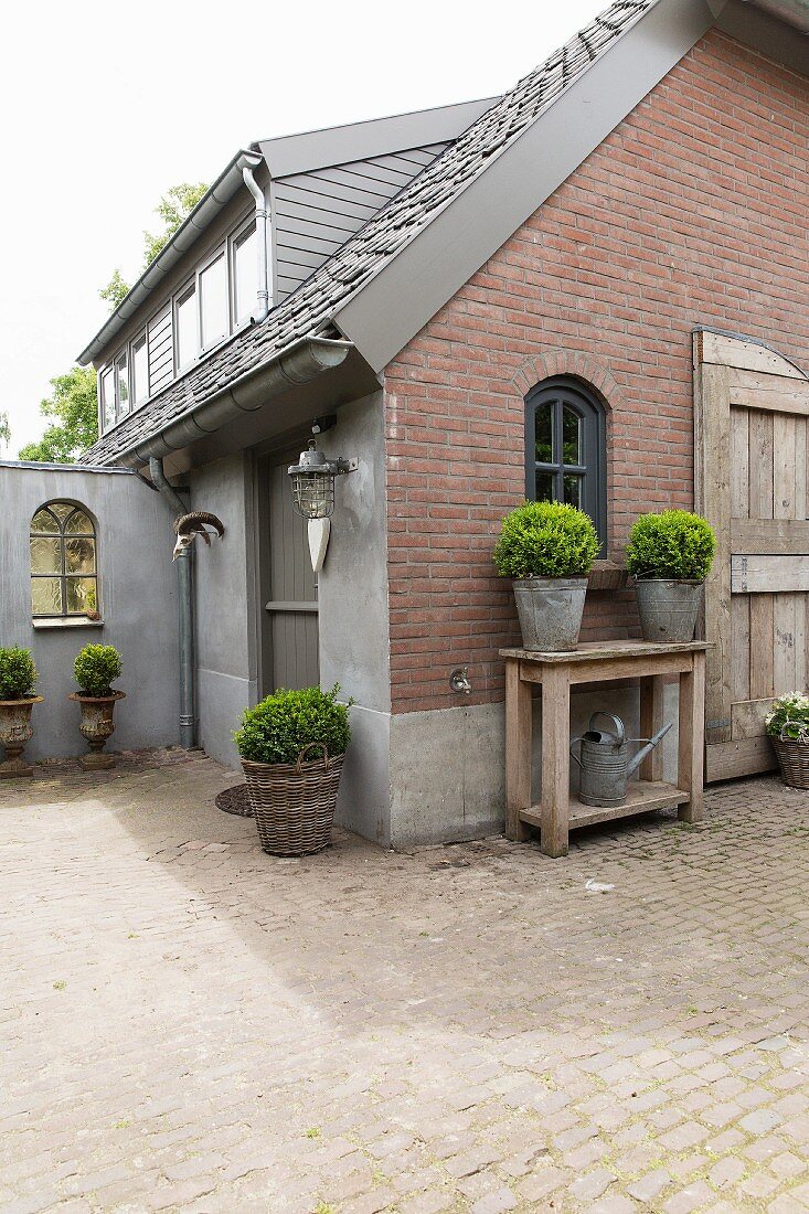 Planters on wooden table against brick gable-end wall and paved courtyard outside house with dormer windows