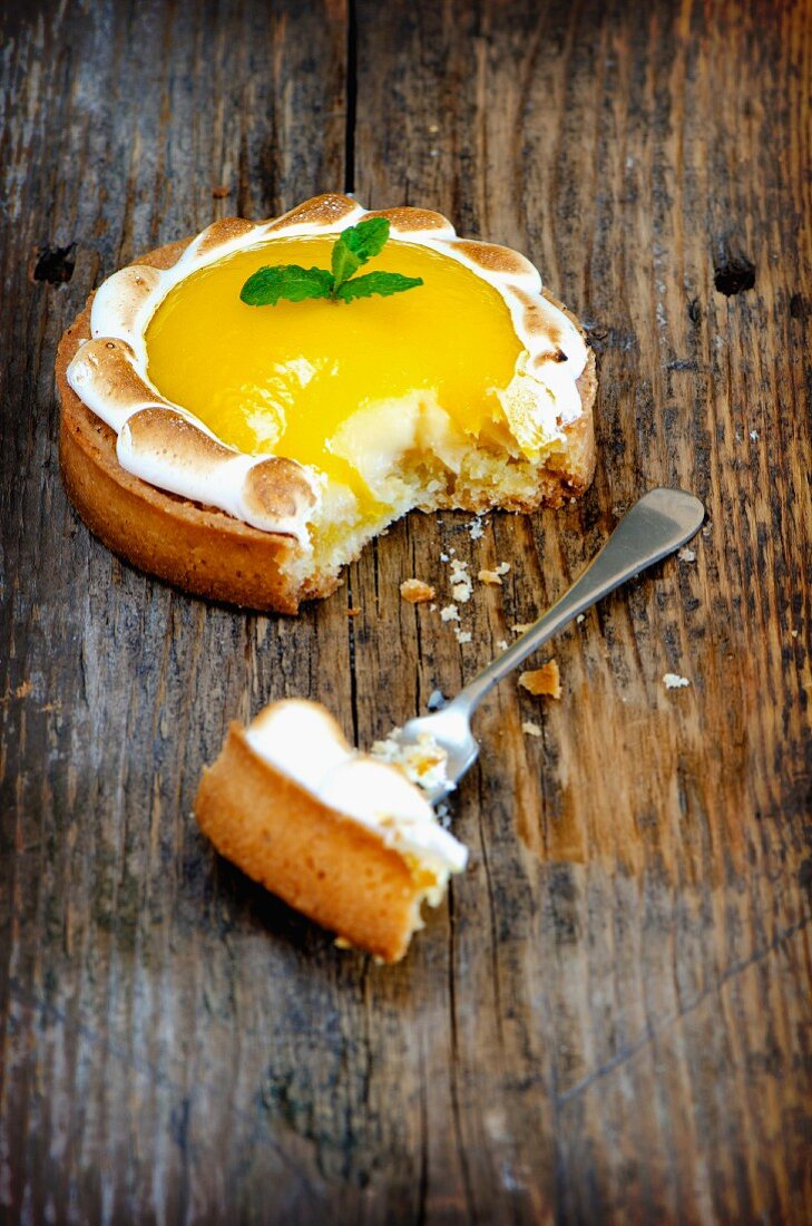 A lemon meringue tartlet on a wooden surface with a bite taken out of it