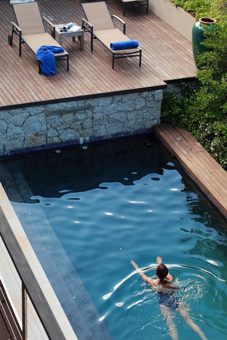 Frau schwimmt in Pool unterhalb eines Holzdecks mit Sonnenliegen
