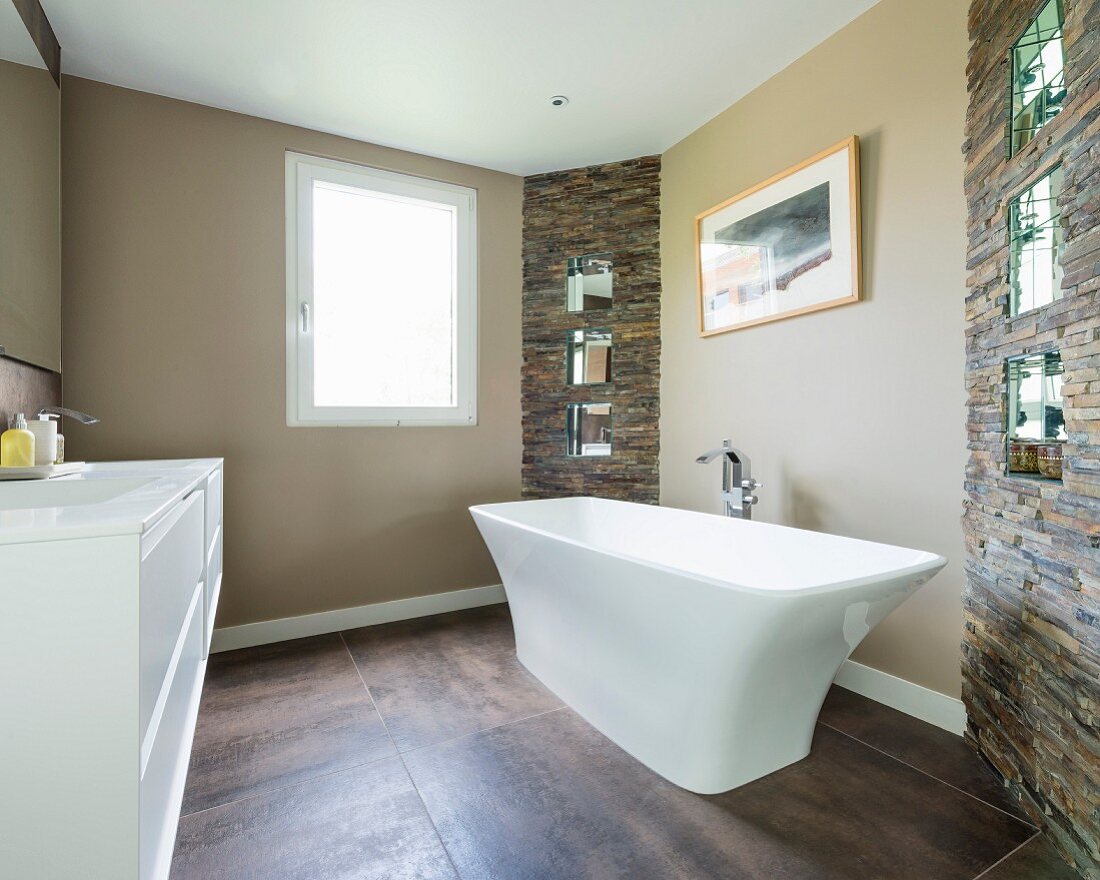 Free-standing bathtub on large floor tiles in modern bathroom with sections of stone wall