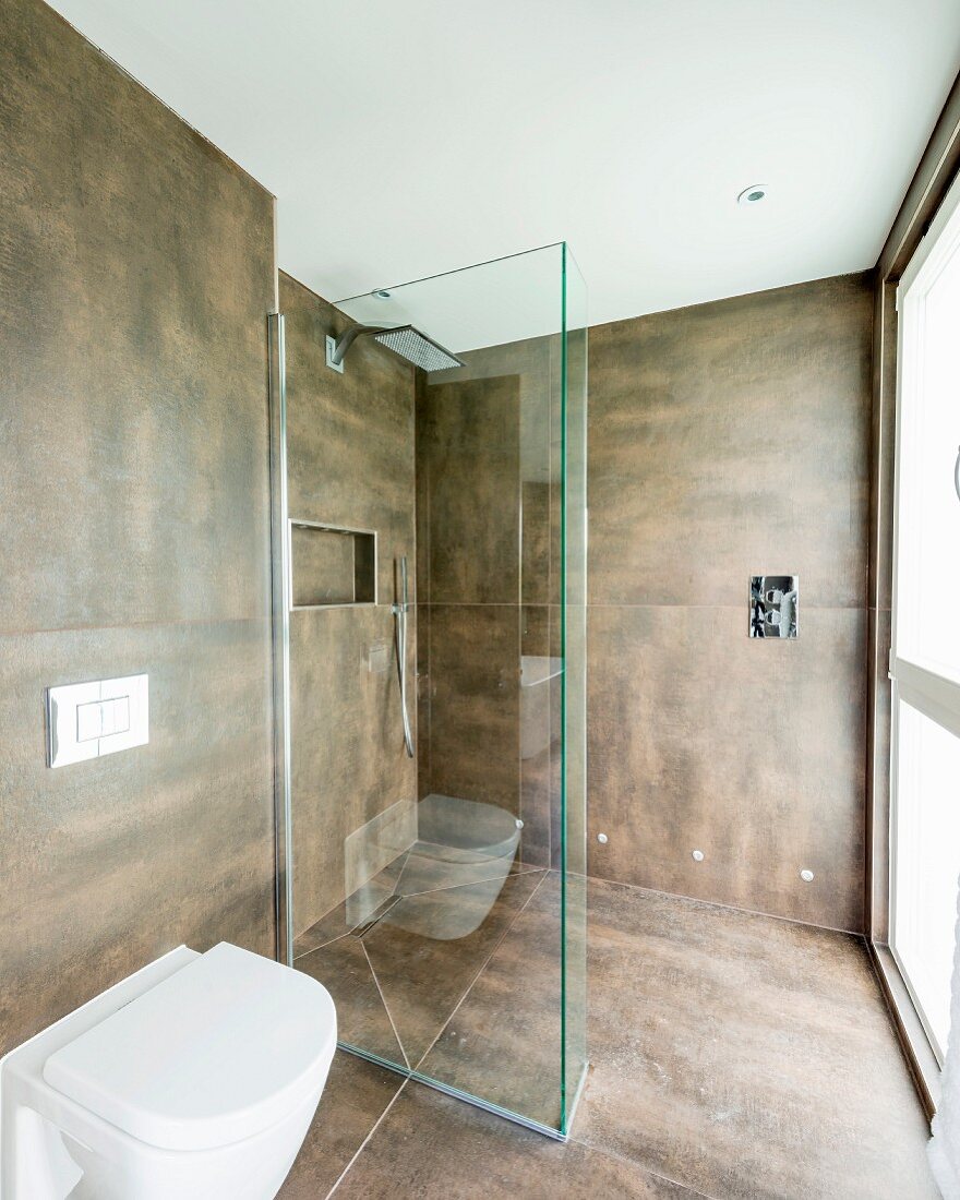 Toilet next to glazed, floor-level shower area in modern bathroom with large brown tiles on walls and floor