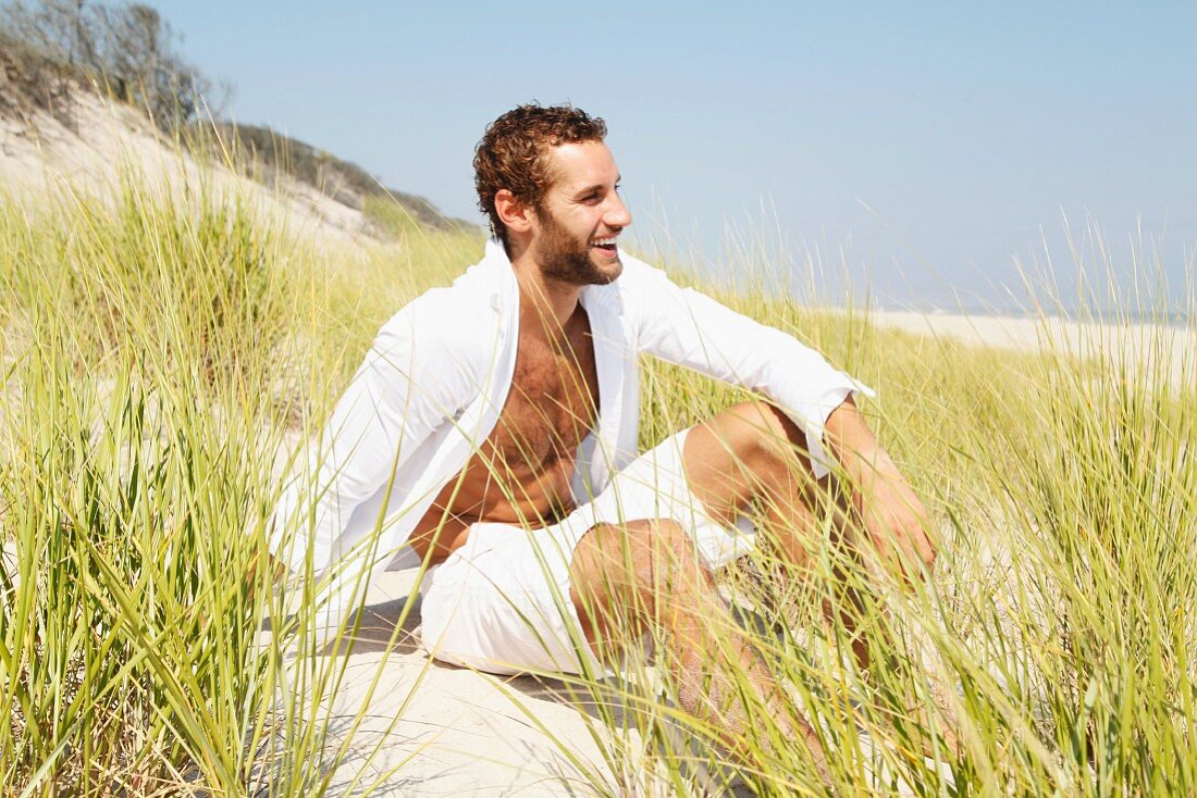 Junger Mann in weisser, luftiger Kleidung sitzt in Sanddüne am Strand