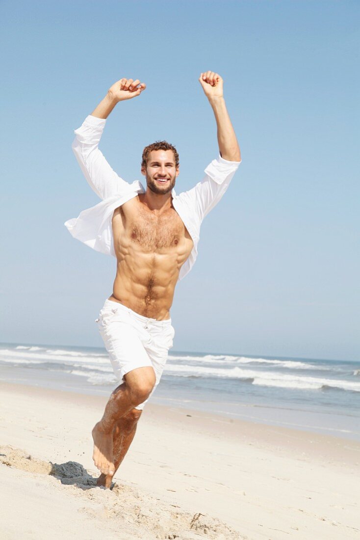 Junger Mann in Shorts und offenem Hemd läuft am Strand
