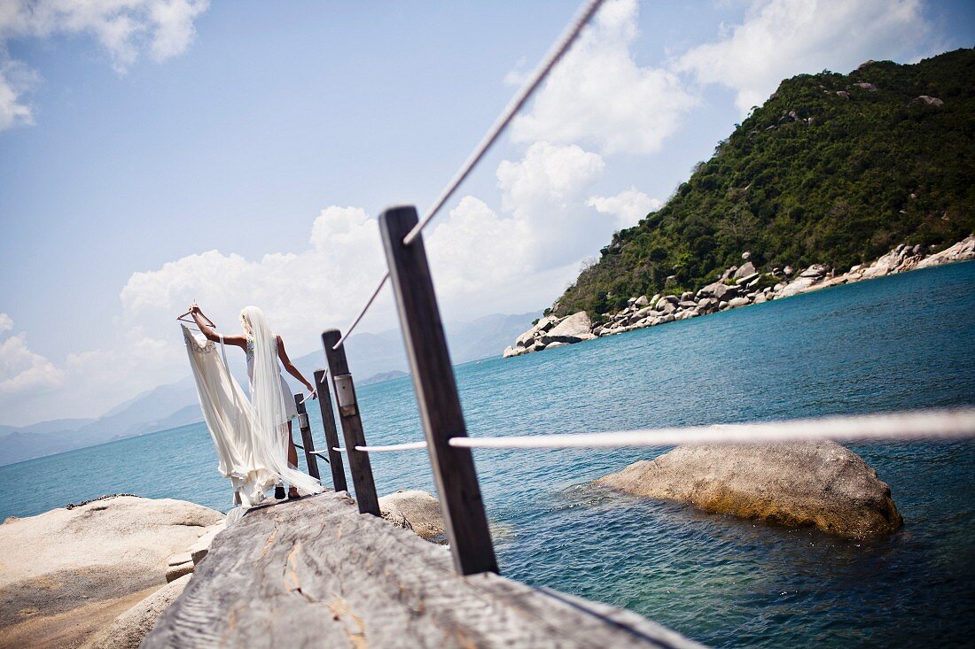 Frau in Unterkleid und Schleier, Brautkleid auf Kleiderbügel haltend, steht auf Steg am Meer (Nha Trang, Vietnam)