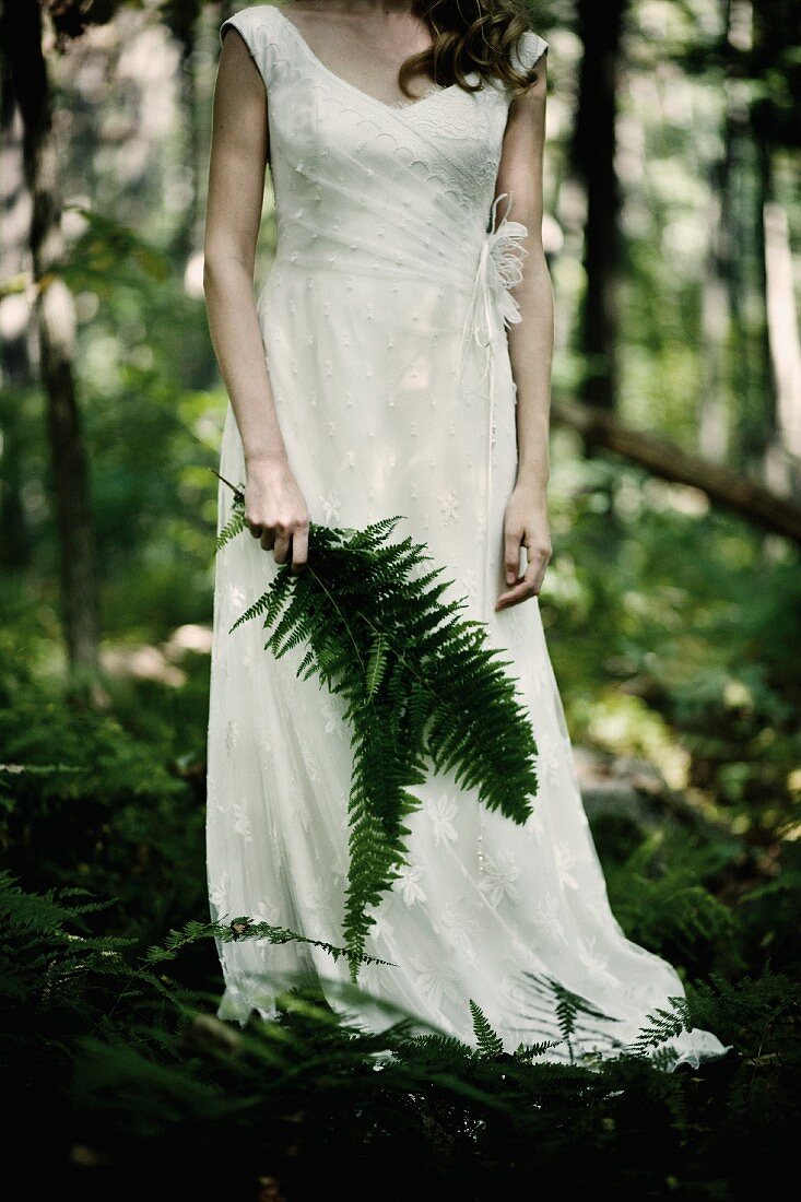 Braut steht im Wald mit Farnblättern in der Hand