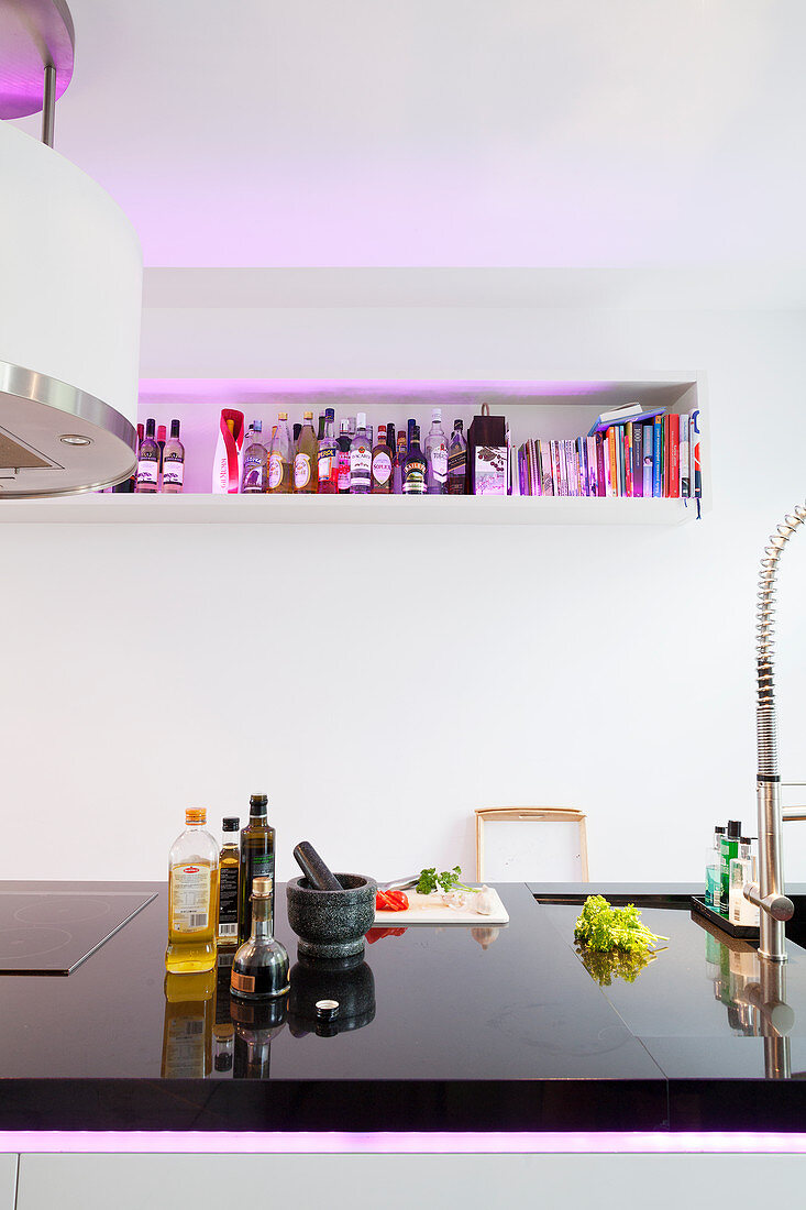Kitchen utensils on black polished worksurface of island counter opposite wall-mounted shelf illuminated by purple light