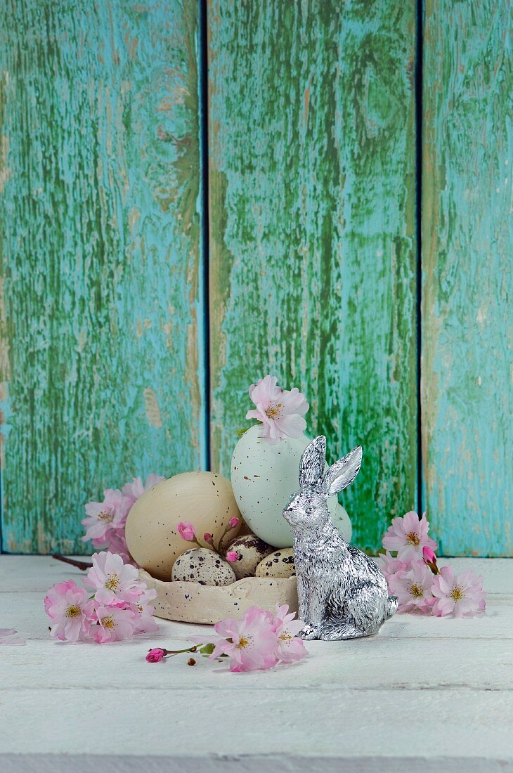 Easter eggs and quail's eggs in bowl next to silver Easter bunny and cherry blossom on wooden surface
