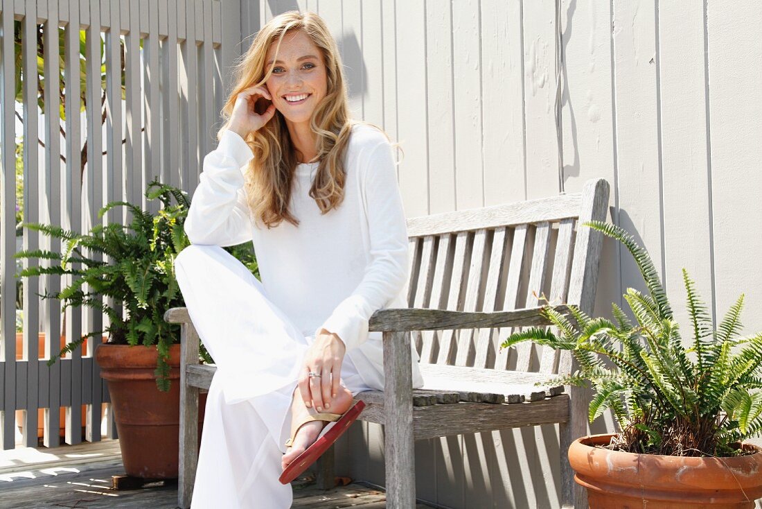 A blonde woman wearing loose-fitting, white clothing sitting on a bench on a terrace