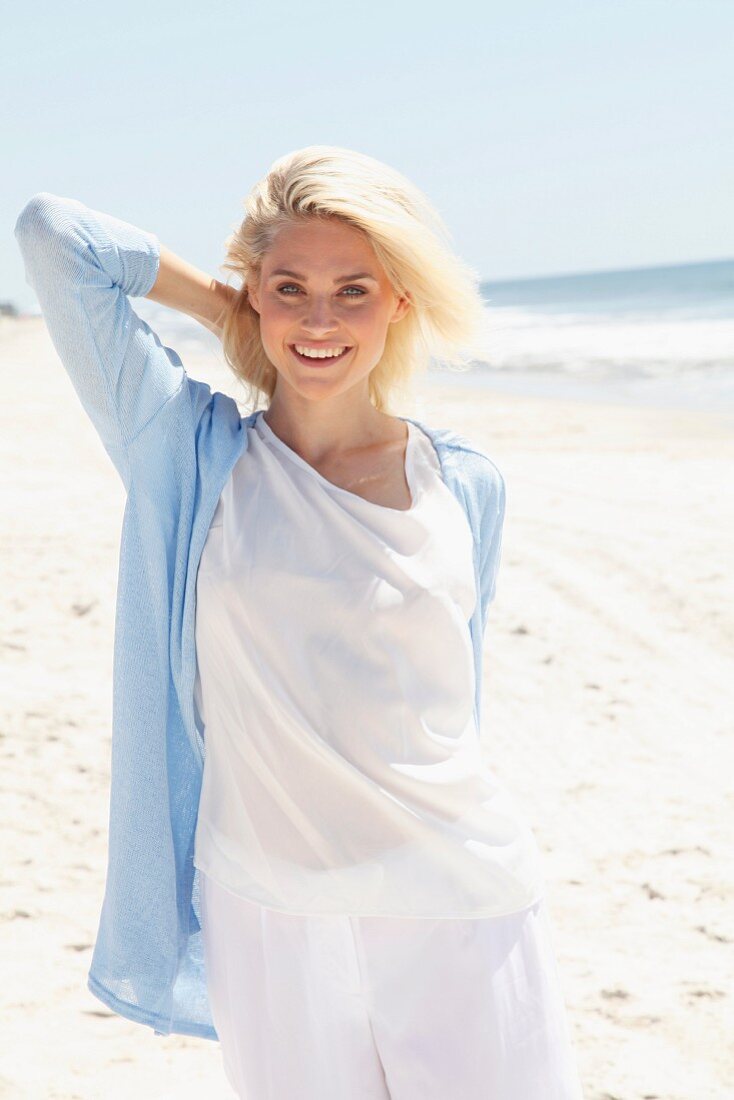 Blonde Frau in weißem Shirt, Hose und hellblauer Strickjacke am Strand