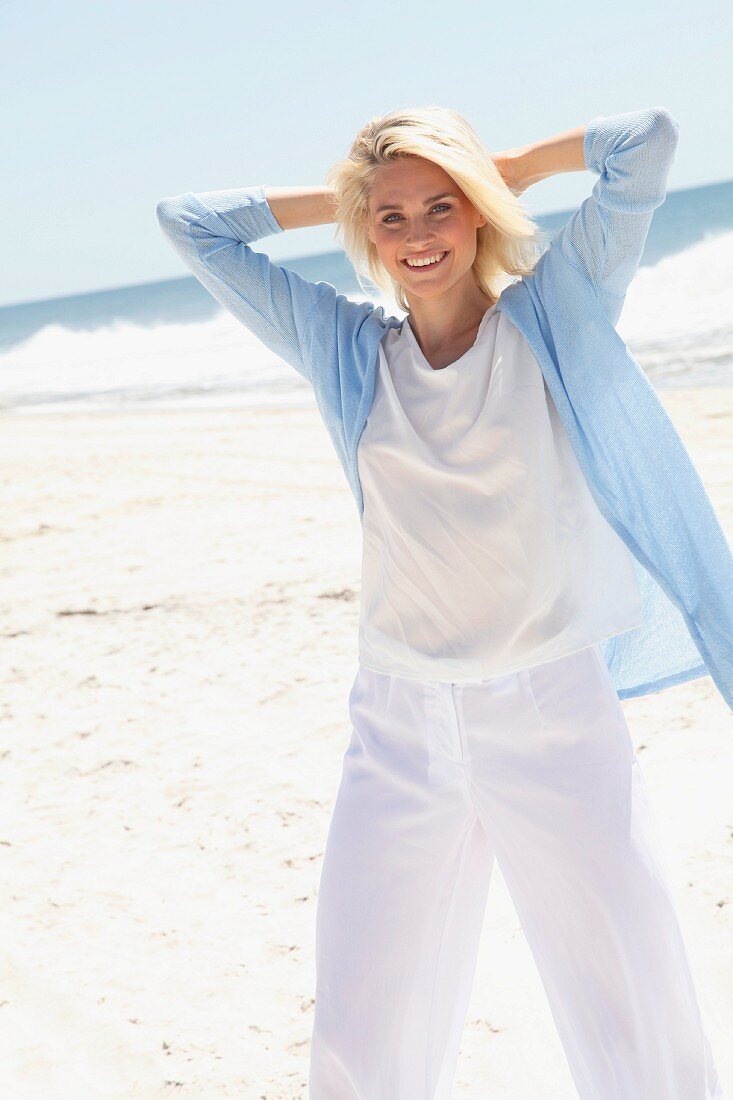 Blonde Frau in weißem Shirt, Hose und hellblauer Strickjacke am Strand