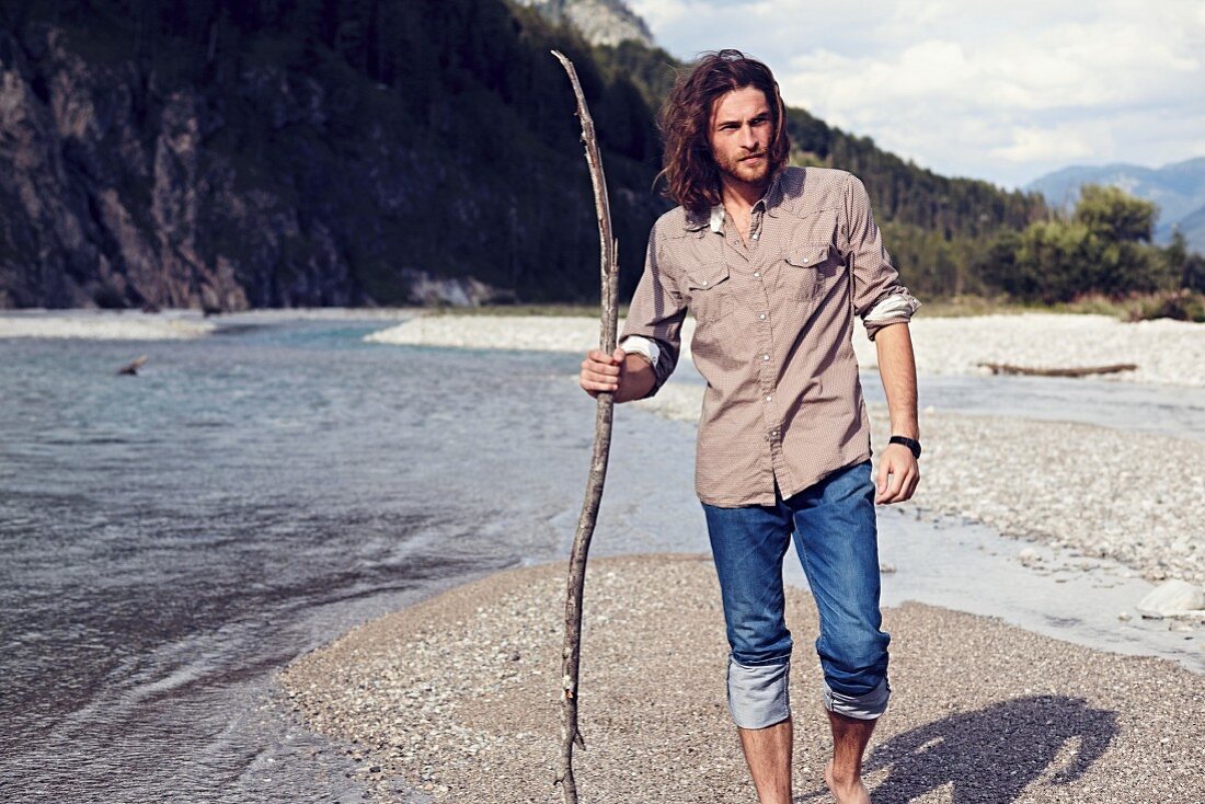 A young man with a beard and long hair walking along a gravelly river bed