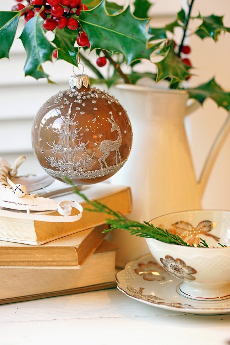 Christmas bauble with forest motif hanging from holly in jug next to stack of books and teacup