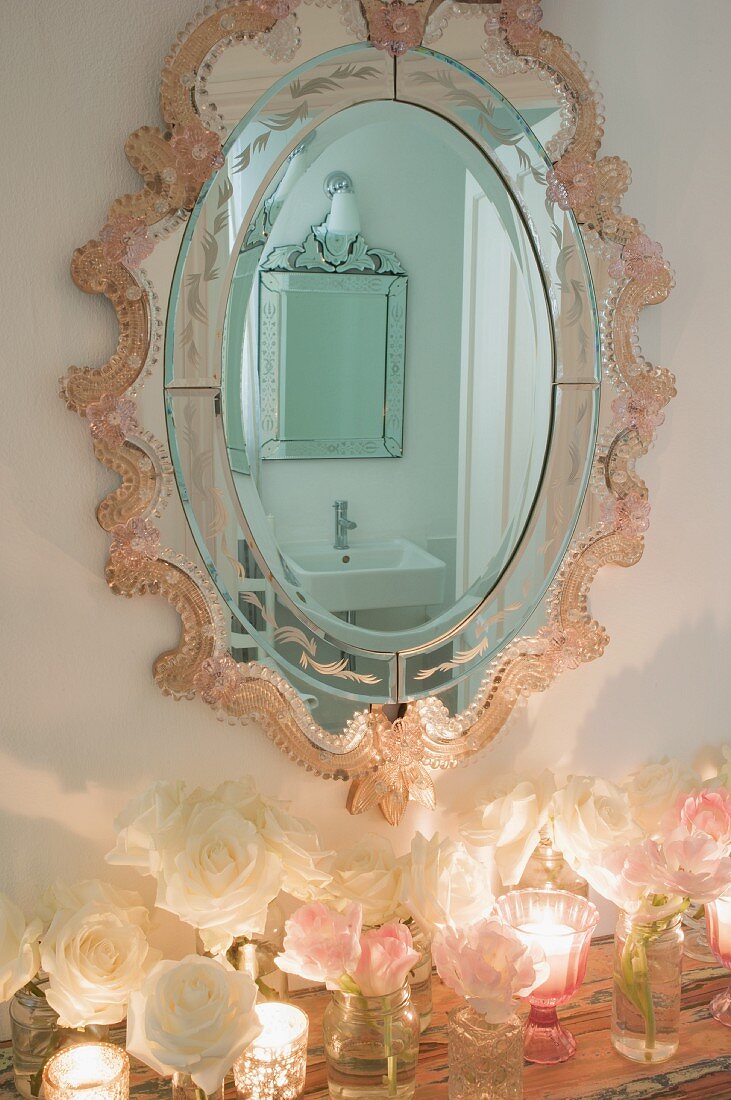 Romantic, ornate oval mirror reflecting bathroom above arrangement of roses and tealight holders