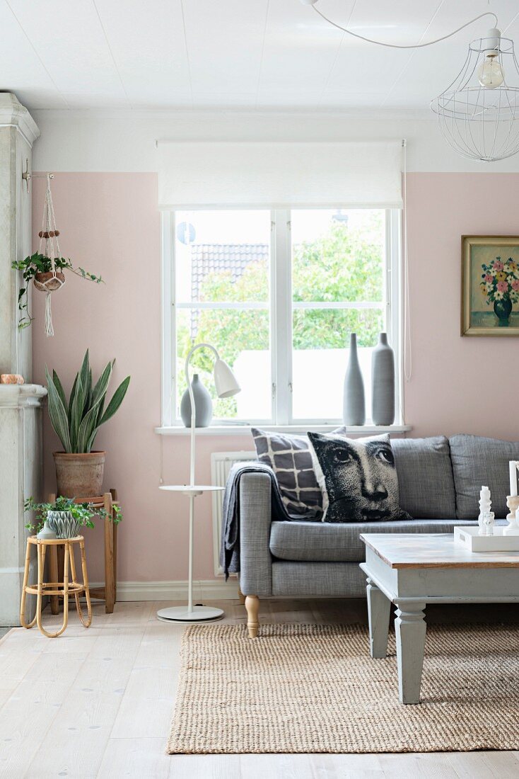 Scatter cushions with face motifs on sofa below window next to potted plants on simple stools in romantic living room with pink-painted walls