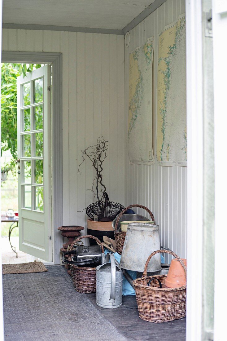 Wicker baskets and gardening utensils on floor of hall with white wood panelling and open garden door in background